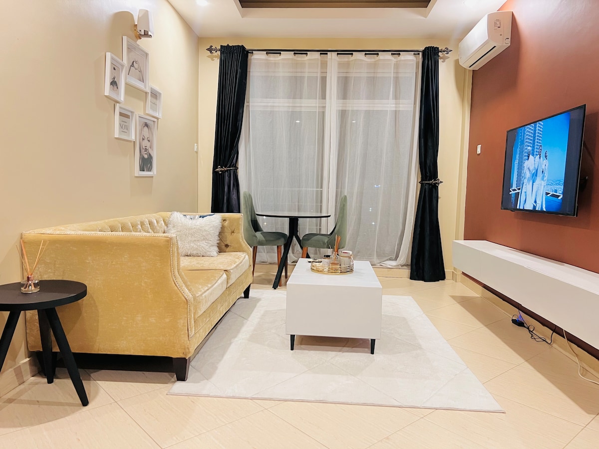 A modern living area is equipped with a plush yellow sofa and a sleek black side table. A small dining table with green chairs is situated nearby. Light filters through sheer curtains, and a television is mounted on a white wall unit opposite the seating area.