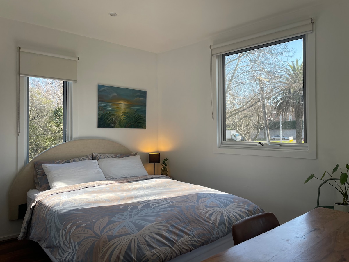 A bright bedroom features a spacious bed with a patterned blanket. Two windows allow natural light to fill the room, while a painting adorns the wall above the bed. A small bedside lamp and a plant add a touch of greenery to the space.