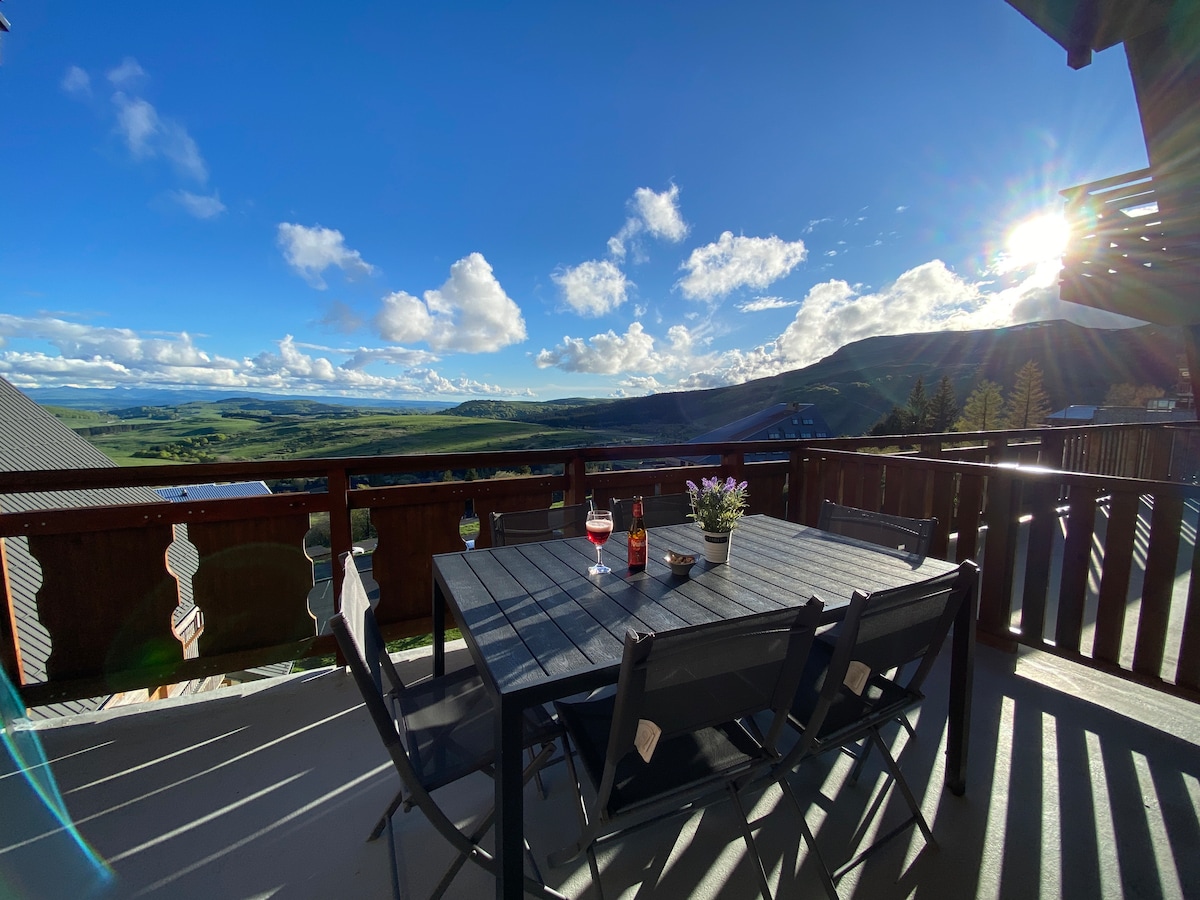 A balcony table is set for six, featuring a bottle of wine and glasses. The expansive view encompasses rolling hills and vibrant skies, illuminated by sunlight. Shadows of the railing and chairs create a pattern on the deck, inviting relaxation in a serene outdoor setting.