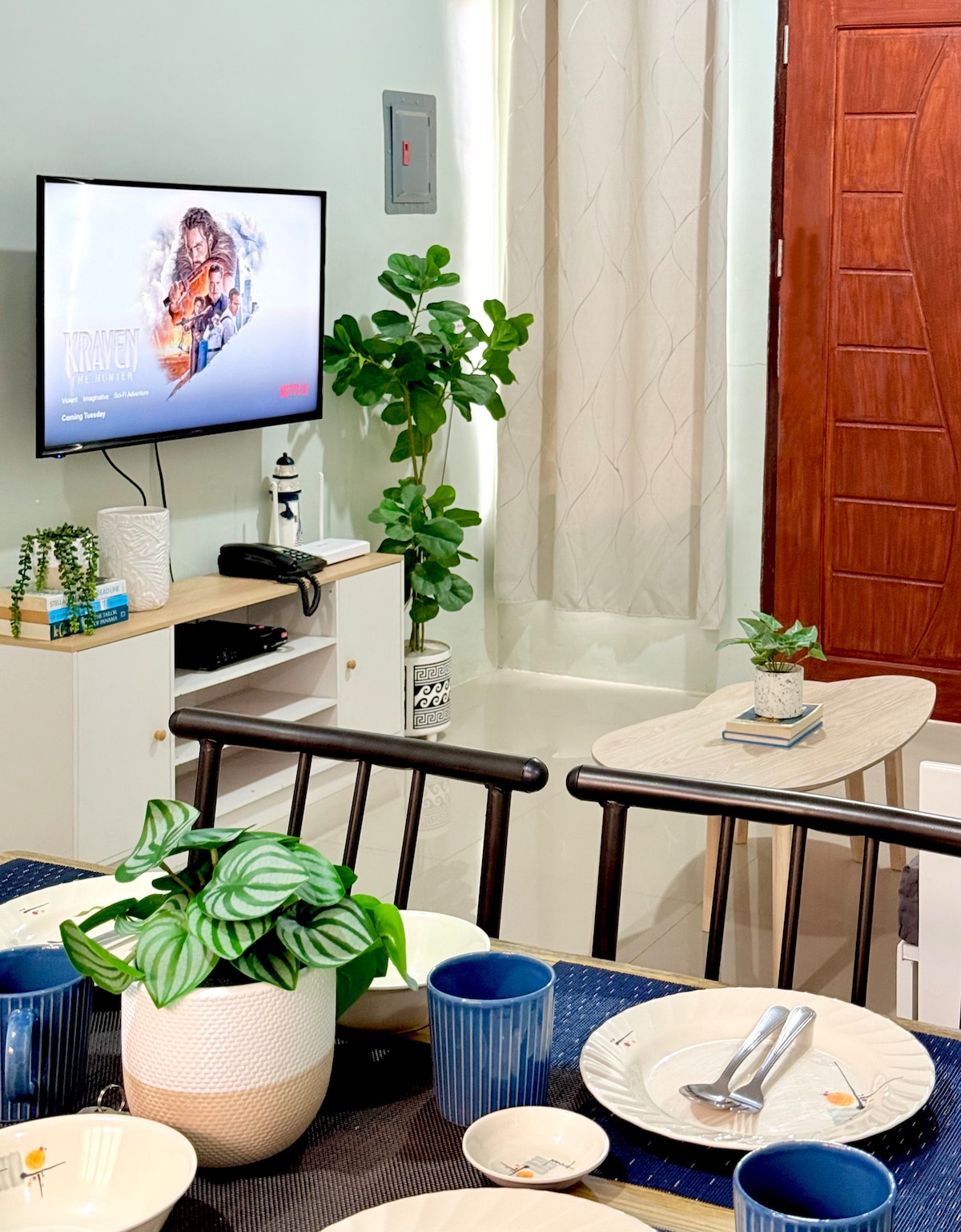 A well-equipped living area features a wall-mounted television displaying a movie, accompanied by a stylish plant near a side table. A dining table is set with plates and glasses, and a decorative plant enhances the inviting ambiance.