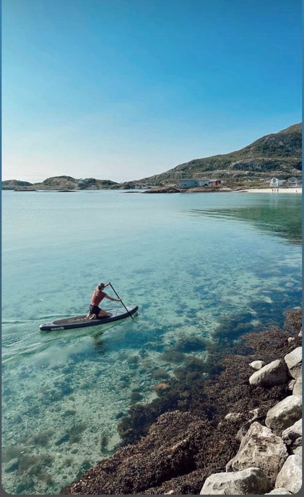 A serene image features a person paddling on a stand-up paddleboard across calm, clear waters. The shoreline is visible with rocky formations and gentle waves lapping at the edges. Scenic mountains and a distant beach set a peaceful backdrop under a bright blue sky.