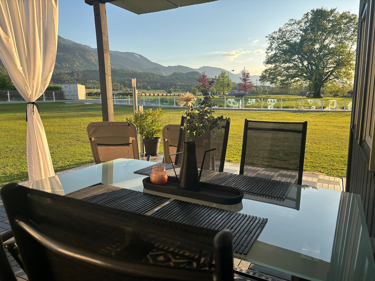 An outdoor dining area is presented with a glass table and four chairs. A central decorative arrangement features a black vase and a candle. The landscape captures a view of mountains and trees, with greenery extending to the horizon.