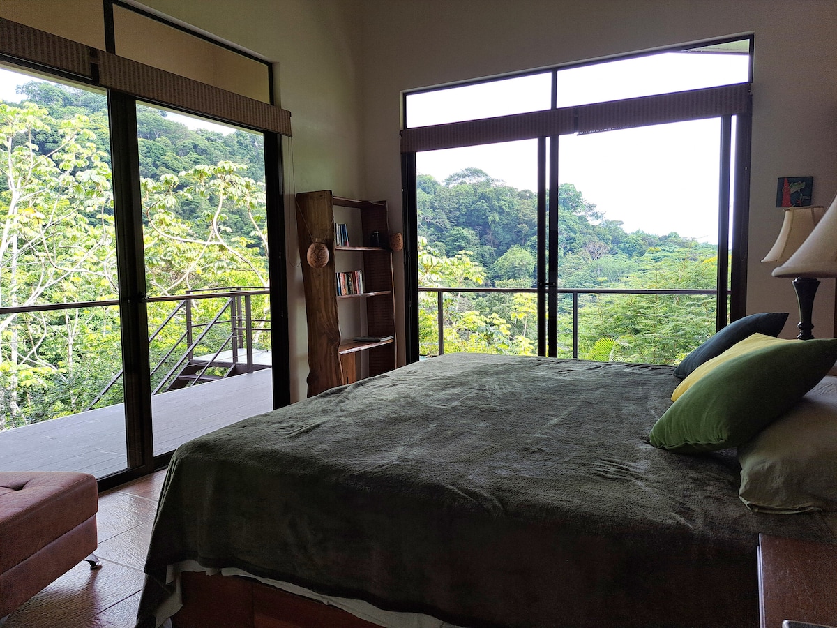 A spacious bedroom features a large bed with dark bedding and colorful accent pillows. Floor-to-ceiling windows offer unobstructed views of the surrounding lush greenery. A cozy seating area and a bookshelf are positioned nearby, contributing to the serene jungle ambiance.
