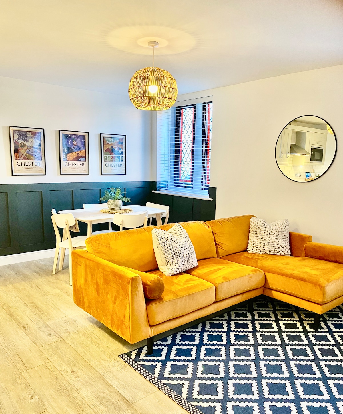 A cozy lounge area features a spacious mustard-colored sofa with decorative cushions. A circular mirror is mounted on the wall, reflecting light from a pendant lamp above. A dining table with white chairs is set against a green wall, adorned with framed artwork.