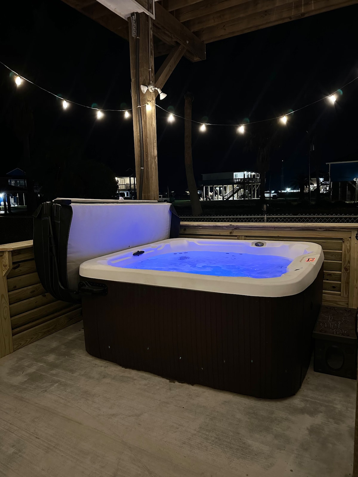 A relaxing hot tub is positioned on a covered deck, illuminated by soft blue lighting. String lights overhead provide a warm glow, and palm trees are visible in the background. The surrounding area features a wooden structure that enhances the outdoor atmosphere.