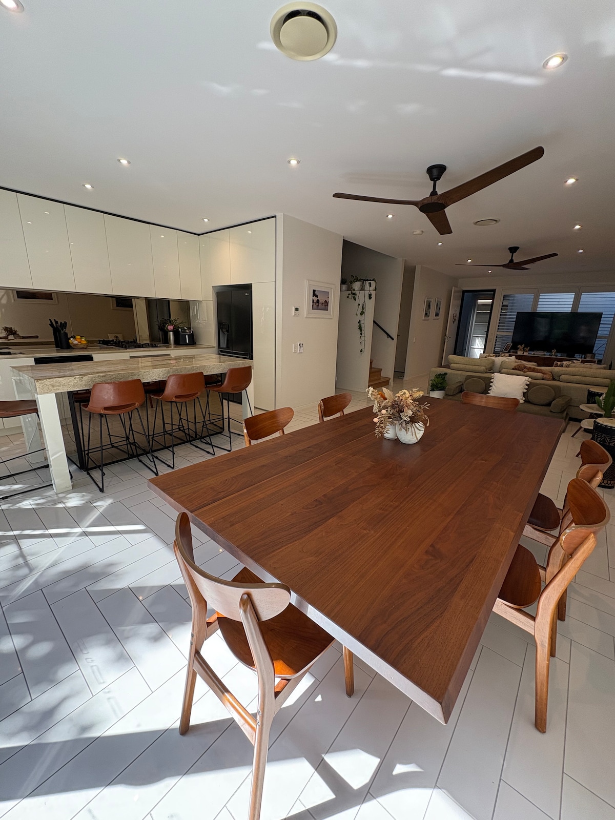 A spacious dining area features a long wooden table surrounded by several wooden chairs. Natural light illuminates the room, which includes a modern kitchen visible in the background. Light-colored tiled flooring complements the warm tones of the furniture.