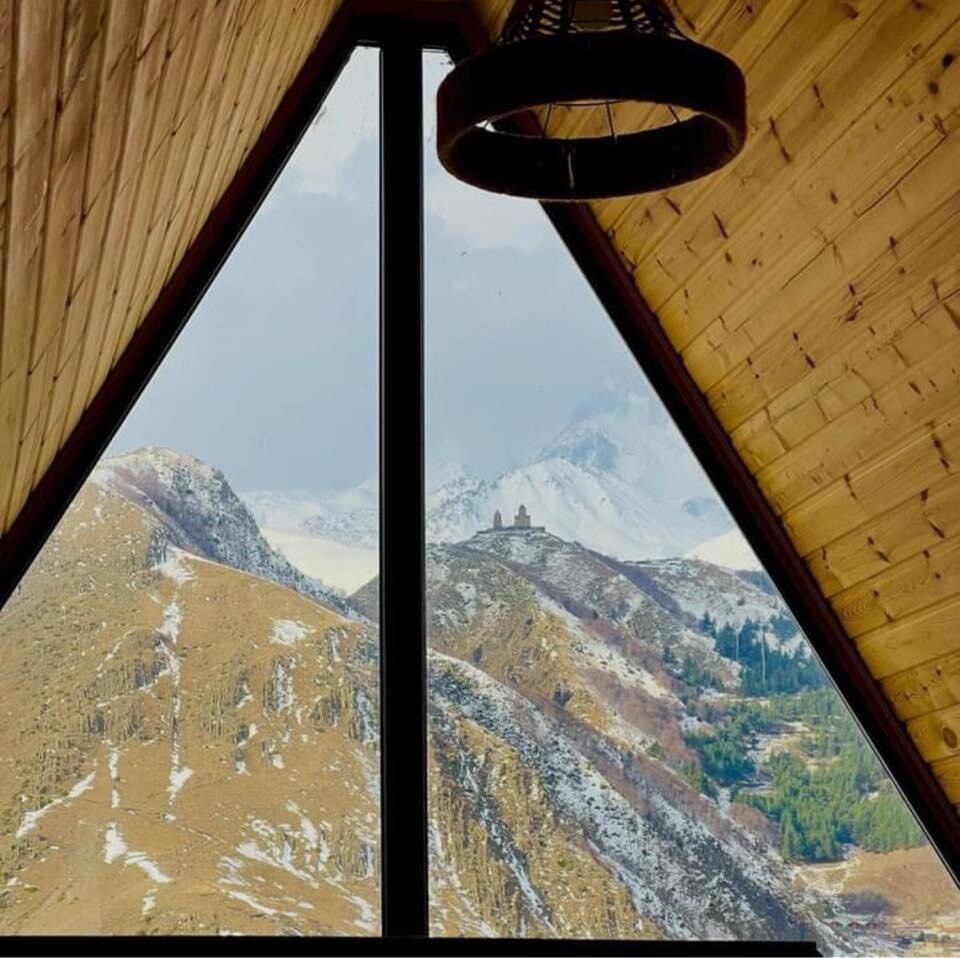 A triangular window frames a breathtaking view of the snowy Mkinvartsveri mountain and Gergeti Trinity Church, set against a backdrop of rugged hills. Natural wood paneling adds warmth to the interior space.