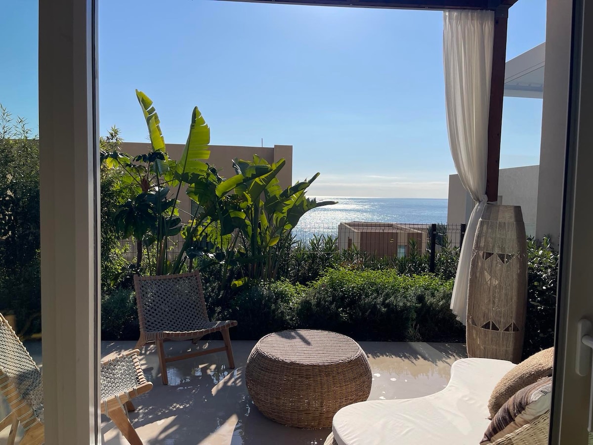 A serene outdoor space is showcased, featuring comfortable lounge chairs and a round woven table. Lush greenery surrounds the area, with large leafed plants in view. The backdrop presents tranquil sea views under a clear blue sky, creating a peaceful atmosphere.