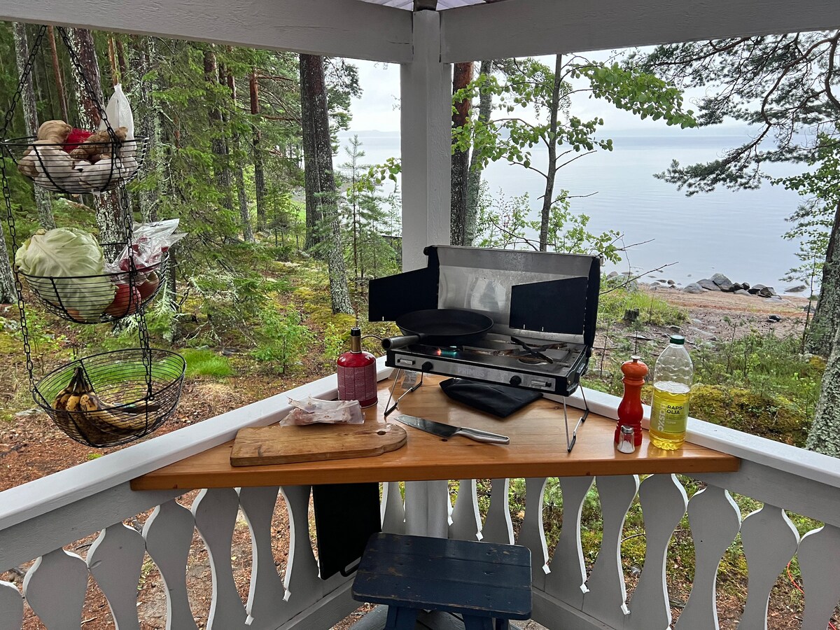 An outdoor cooking area is presented, featuring a compact gas stove and a wooden countertop. A collection of condiments and cooking supplies is visible, alongside hanging baskets filled with fresh produce. Surrounding trees and a view of the tranquil lake create a serene atmosphere.