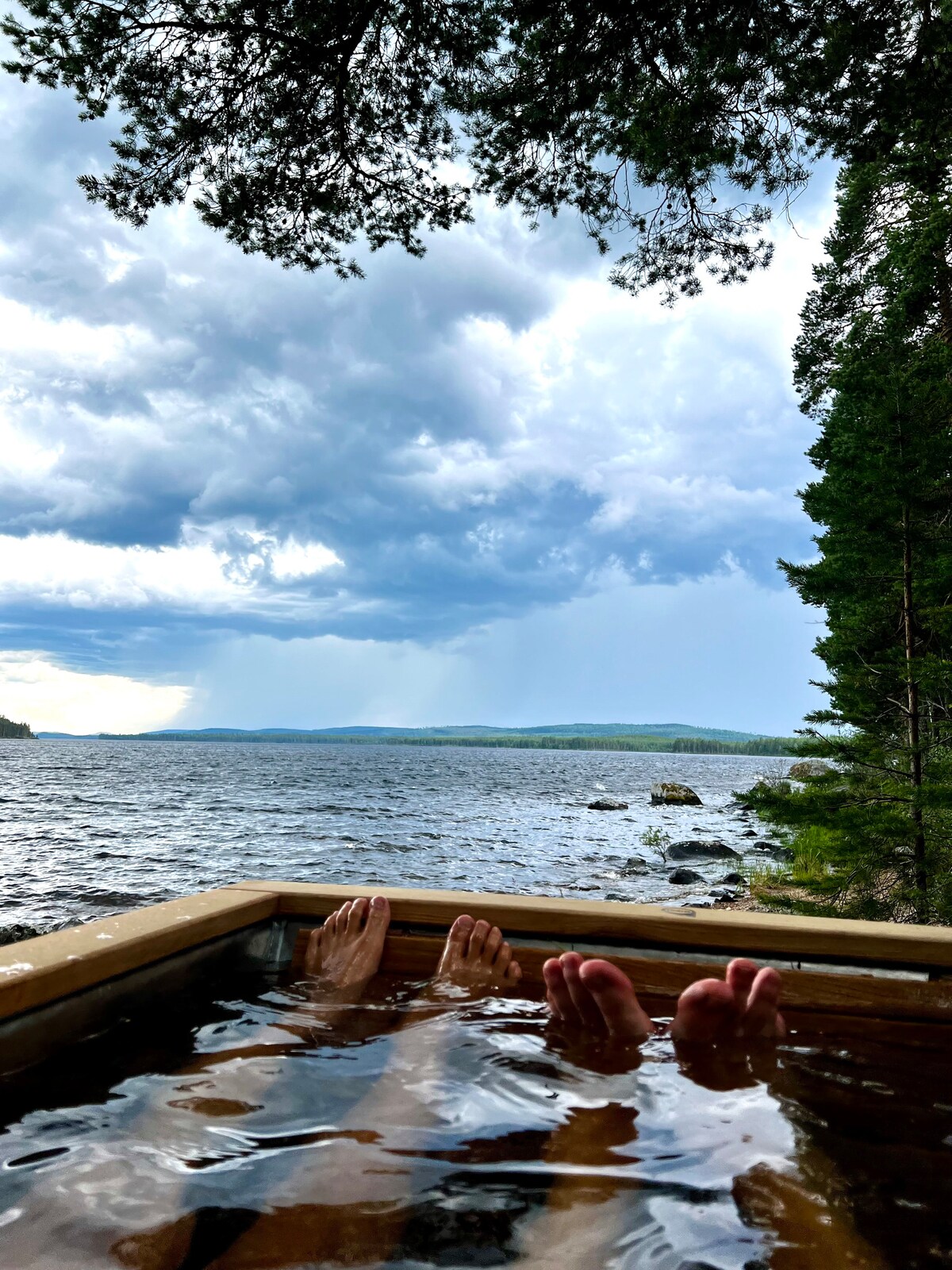 A wooden hot tub sits at the water's edge, with feet resting in the warm water. A scenic view of a lake and distant forest can be seen, under a dramatic sky with dark clouds. The shoreline is lined with rocks, enhancing the natural setting.