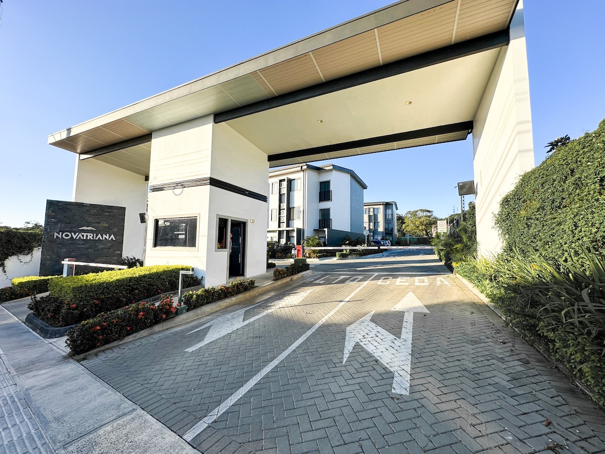 The entrance of a gated condominium is depicted, featuring a modern structure with a prominent sign displaying 'NOVATRIANA.' Neatly landscaped green areas with colorful plants border the drive, and clear arrows indicate the path for vehicles entering. A bright blue sky is visible above.
