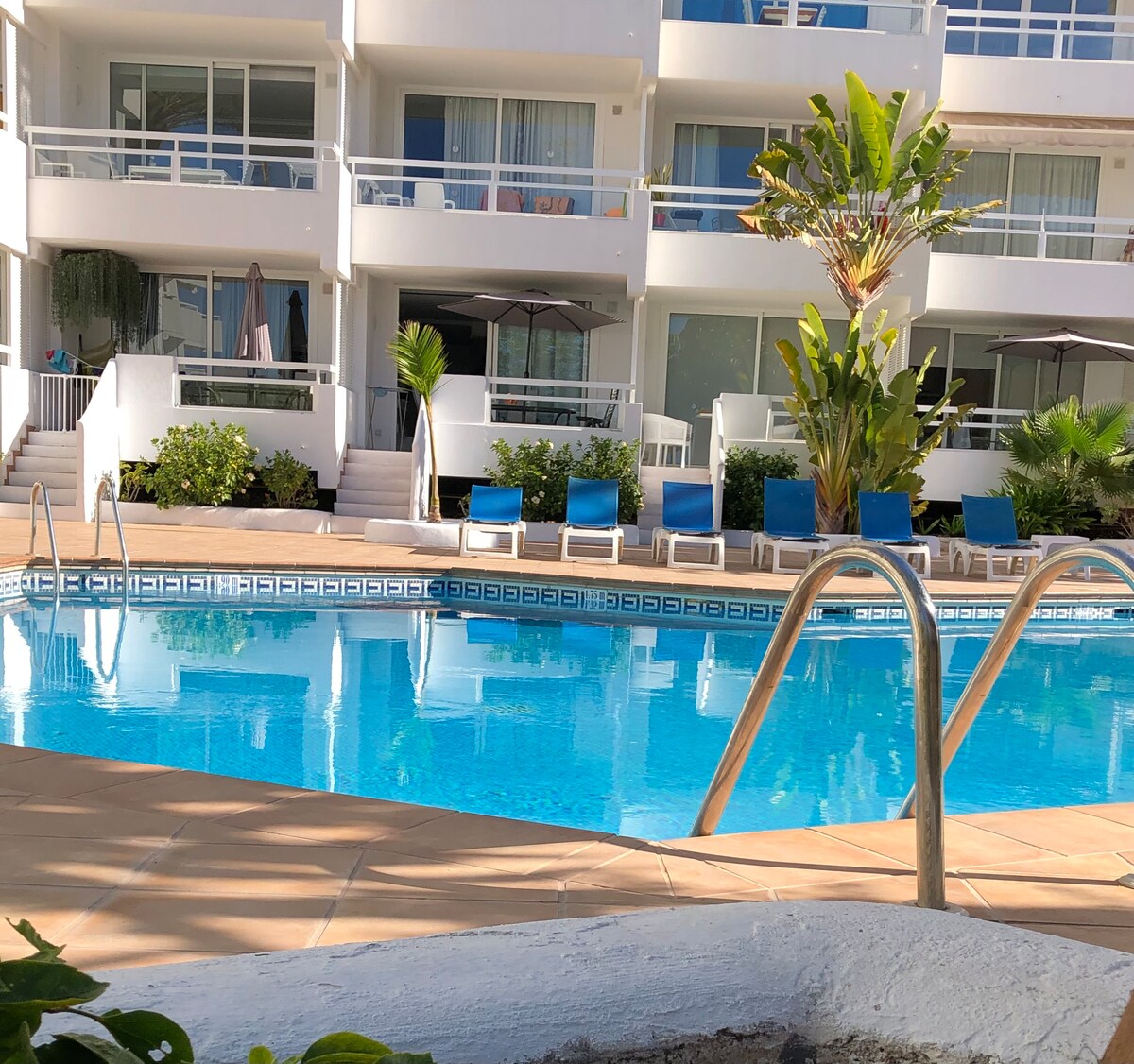 A clear swimming pool is surrounded by lounge chairs with blue cushions, creating a relaxing outdoor space. The pool reflects the sunlight, with nearby tropical plants adding greenery. The multi-story building in the background features balconies and large windows, enhancing the open atmosphere.