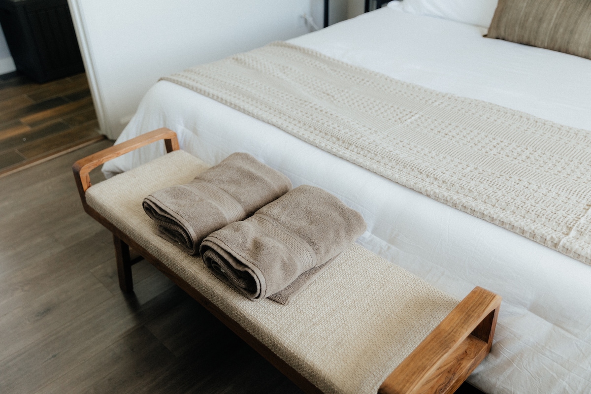 A cozy sleeping area is highlighted by a spacious bed dressed in crisp white linens. On a wooden bench at the foot of the bed, two neatly rolled towels are arranged, adding a touch of comfort to the serene setting.