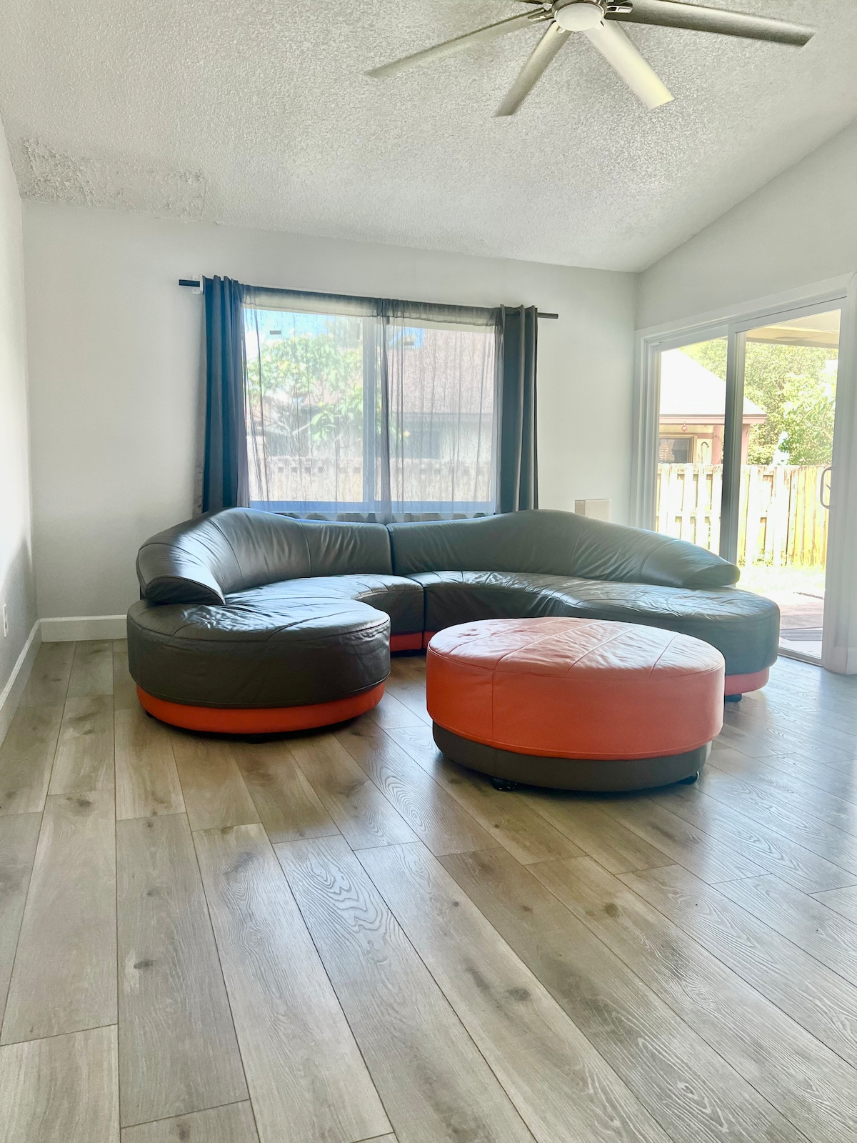 A spacious living area features a large circular sectional sofa in black and orange hues, complemented by matching ottomans. Natural light streams through expansive windows, illuminating the wood-look flooring. Sliding glass doors provide access to the fenced backyard.