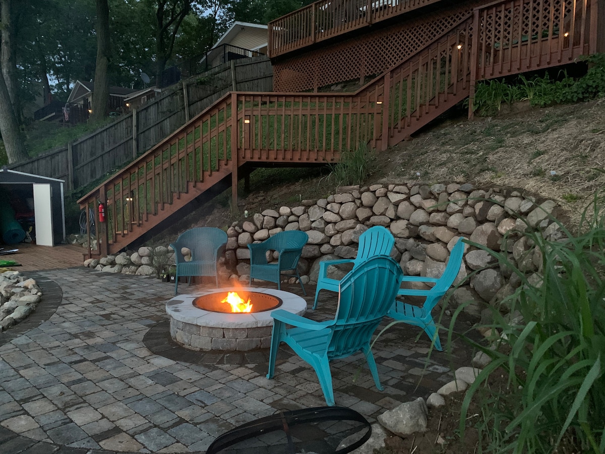A cozy lakeside patio features a circular fire pit with flames flickering. Four teal adirondack chairs are arranged around the fire, set on a stone patio. A sloping staircase leads up to a wooden deck, surrounded by landscaped stones and greenery.
