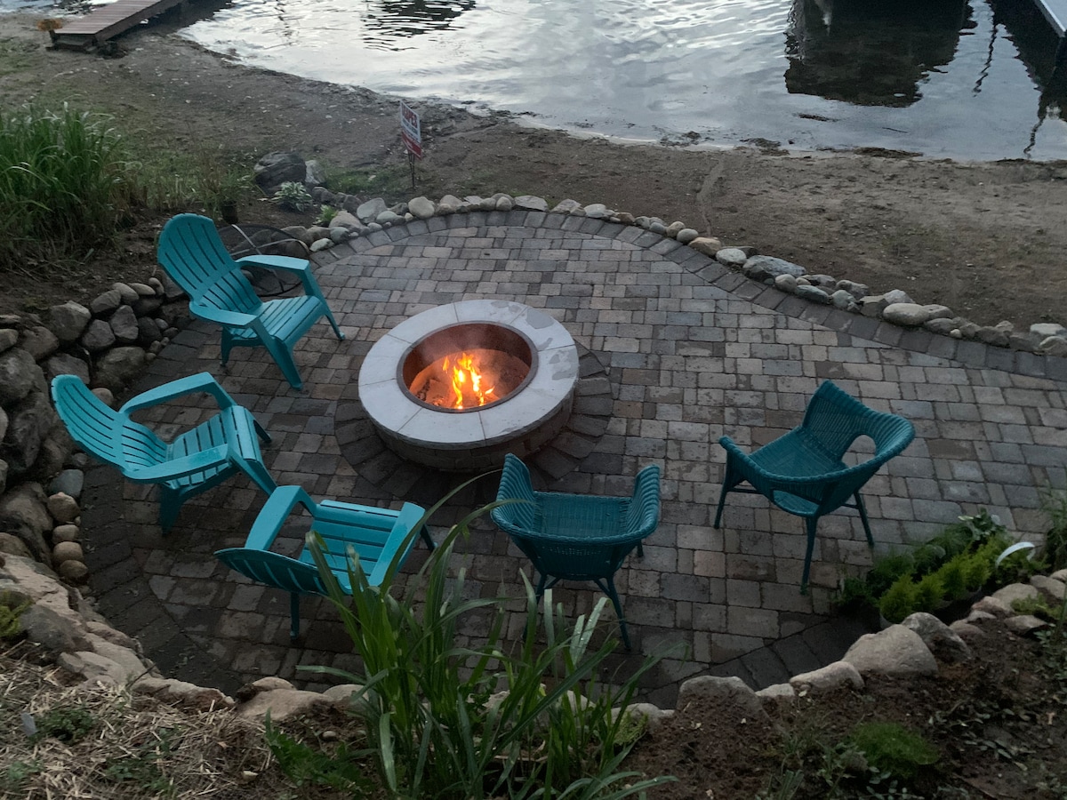 A circular stone fire pit is surrounded by a patio of pavers, featuring five teal-colored chairs arranged for seating. The area is bordered by natural stone and greenery, with the nearby water reflecting soft evening light.