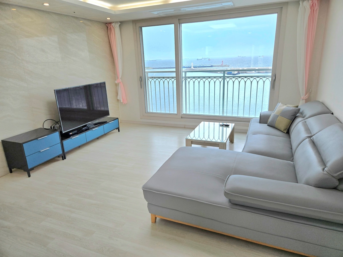 A light-filled living area features large floor-to-ceiling windows with a view of the water. A comfortable gray sectional sofa is positioned facing a modern television, while a glass coffee table sits in the center of the space, enhancing the room's open feel.