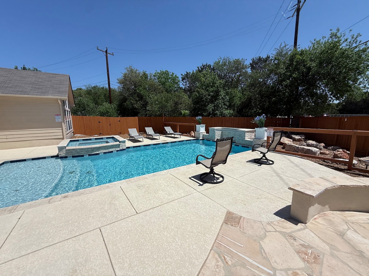 A spacious outdoor area features a newly built inground pool, surrounded by a concrete deck. Several lounge chairs are positioned along the pool's edge, with greenery visible in the background. A spa area is situated adjacent to the pool, creating a relaxing atmosphere.
