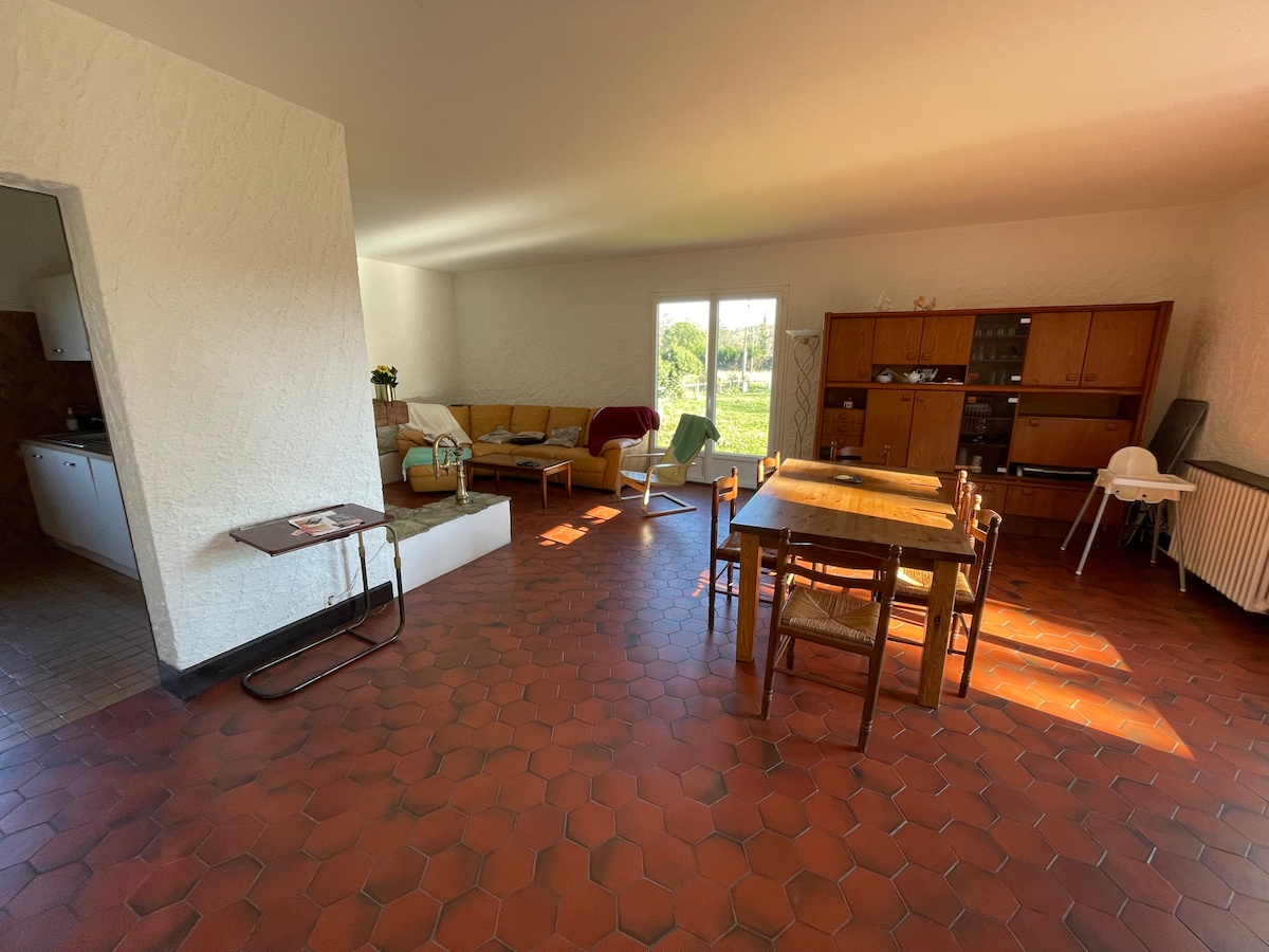 A spacious living area features a dining table surrounded by chairs, illuminated by natural light from large glass doors. A comfortable seating arrangement is visible with a sofa nearby. Textured tile flooring adds warmth to the neutral-toned walls.