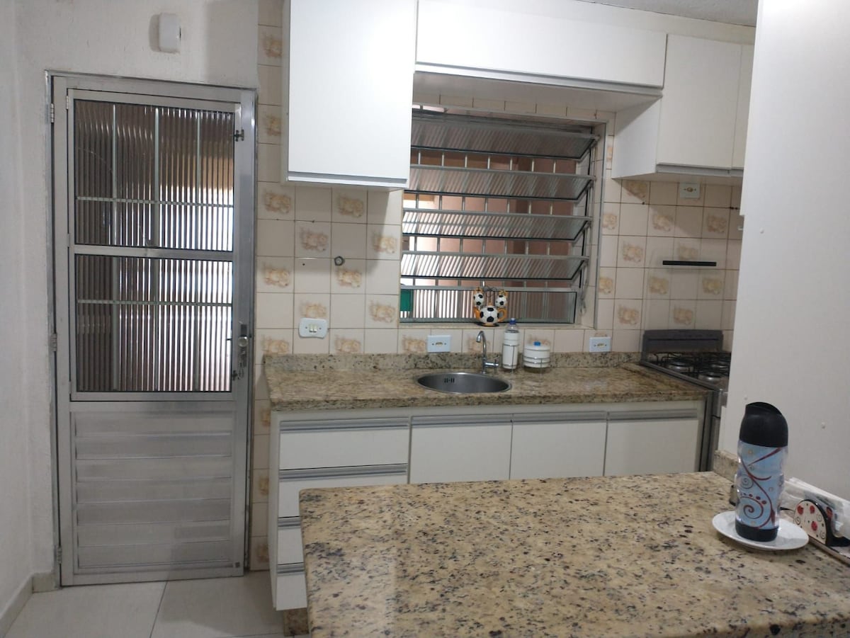 A modern kitchen features light-colored cabinetry and a spacious granite countertop. Stainless steel appliances, including a stove and sink, are visible. Natural light filters through a window adorned with metal bars, while a glass door leads to an adjacent area.