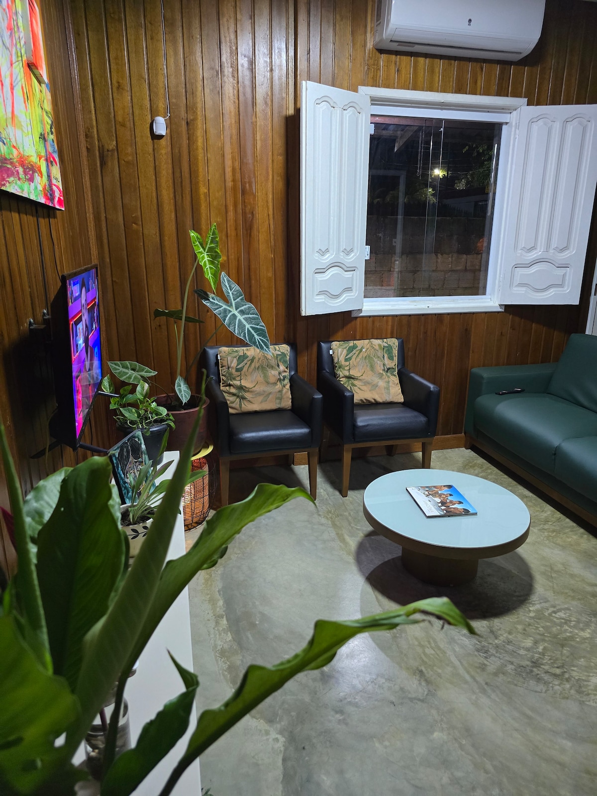 A cozy sitting area is featured with two black armchairs adorned with tropical-patterned cushions. A round wooden coffee table is placed at the center, holding a magazine. Lush green plants are positioned throughout, while a modern television is mounted against a wooden wall.