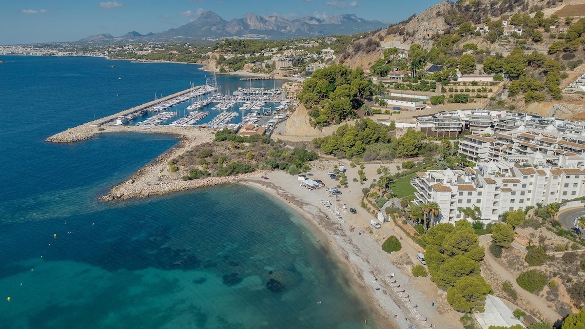 An aerial view reveals a serene coastal area with a sandy beach gently curving along the shoreline. A marina filled with boats is visible, alongside residential buildings set against lush greenery. The surrounding hills provide a scenic backdrop to the tranquil waters.