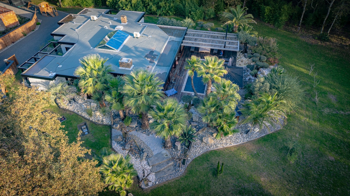 An aerial view of the villa is presented, surrounded by lush greenery and palms. A large terrace is visible, alongside a pool and various landscaped areas featuring rocks and pathways that invite exploration. The serene environment enhances the tranquil ambiance of the property.