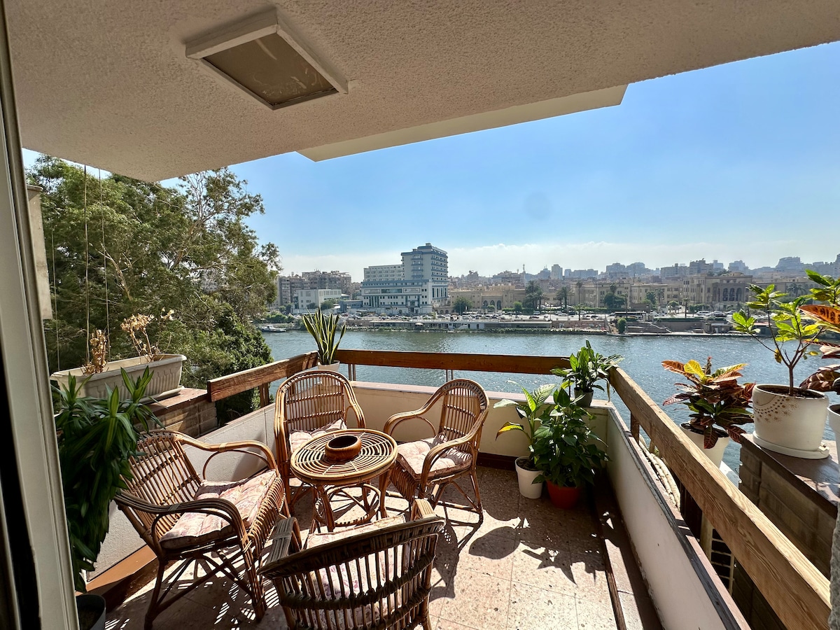 A spacious terrace offers a panoramic view of the Nile, surrounded by potted plants. Several wicker chairs and a circular table are arranged for seating, creating an inviting outdoor space for relaxation and enjoying the scenery.