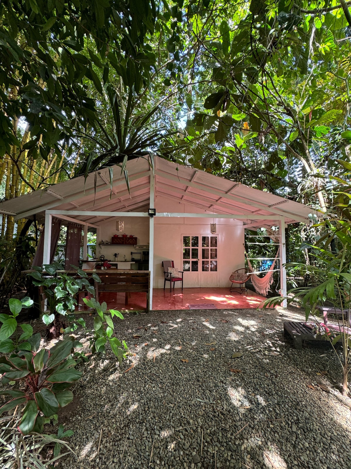 A charming casita is nestled within lush tropical gardens, featuring an open design with a covered porch. A hammock hangs in the corner, while natural light filters through surrounding greenery. The cozy interior is visible through multiple windows, enhancing the connection to nature.
