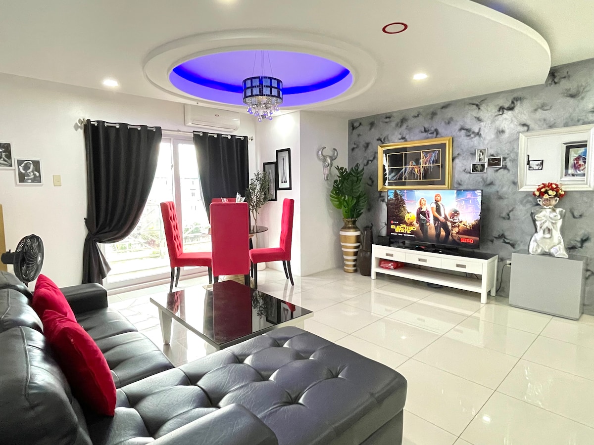 A modern living space features a spacious L-shaped sofa adorned with red and black cushions. A sleek glass coffee table sits centrally, while a smart TV is mounted on the wall, displaying colorful content. A round dining table with red chairs is positioned near large windows, providing natural light.