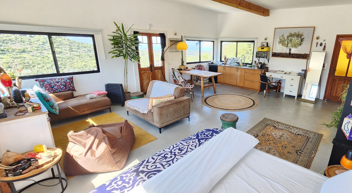 A bright and open loft interior features large windows offering views of the surrounding nature. A comfortable seating area includes a sofa and a colorful armchair. A dining table and workspace are visible, along with a cozy sleeping area and various decorative elements.