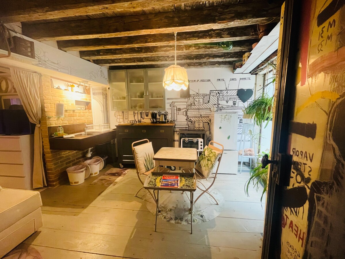 An open-concept living area features a rustic design with exposed wooden beams. A small dining table with two chairs sits near the kitchen, which is equipped with modern appliances. Light fixtures and decorative elements add character to the inviting space.