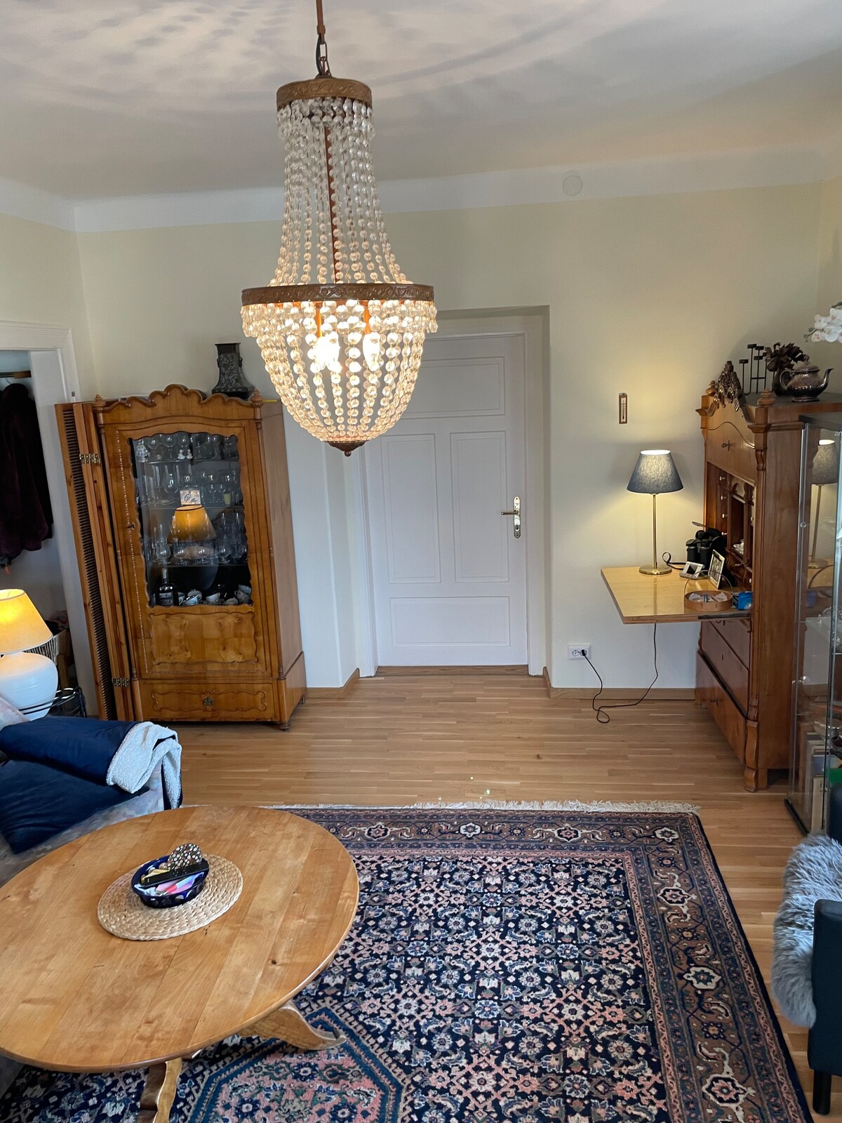 A spacious living area is illuminated by a large chandelier made of crystal beads. A round wooden coffee table sits in the middle, surrounded by soft seating. An antique cupboard and decorative lamps are positioned against the walls, adding warmth to the room.