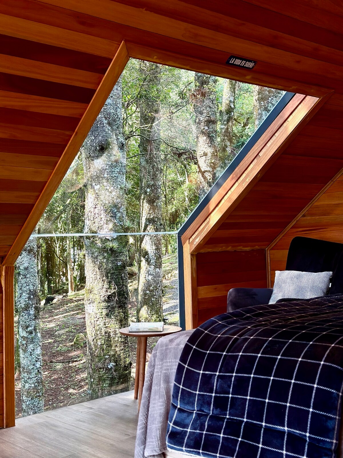 A cozy corner of a cabin features a spacious bed adorned with a soft blanket. Large triangular windows provide a view of the surrounding trees, inviting natural light into the warm wooden interior. A small side table is positioned near the bed, suggesting a peaceful retreat.