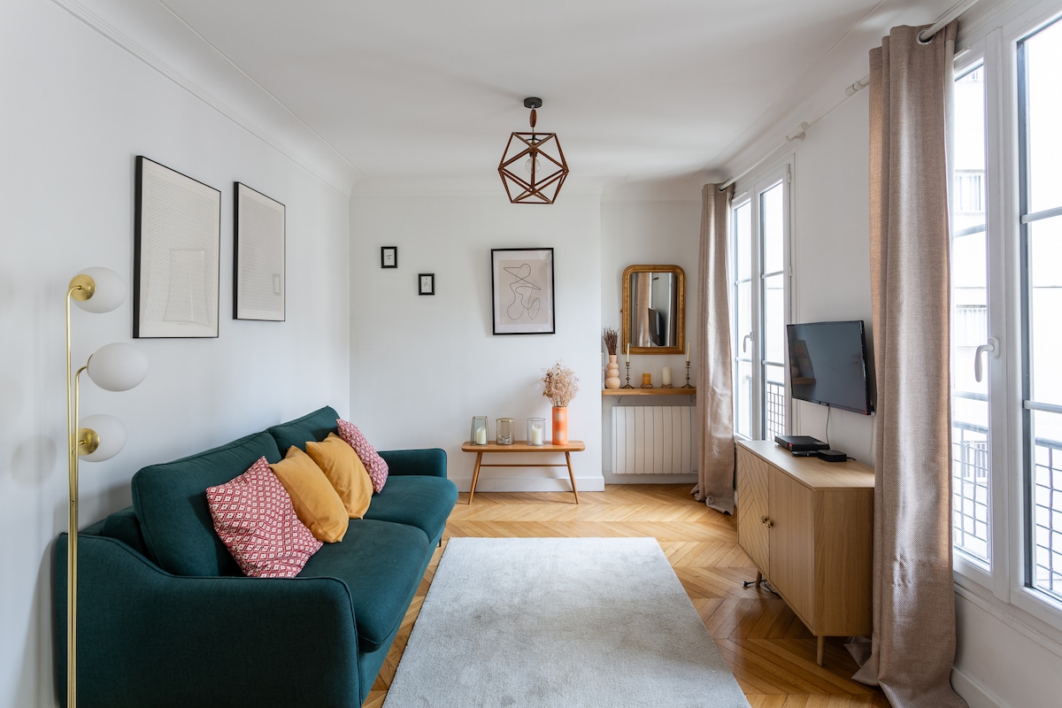 A bright living room is showcased with large windows allowing natural light. A dark green sofa, adorned with colorful cushions, faces a light wood TV stand. A decorative mirror and simple wall art complement the space, which features a soft area rug on light hardwood flooring.