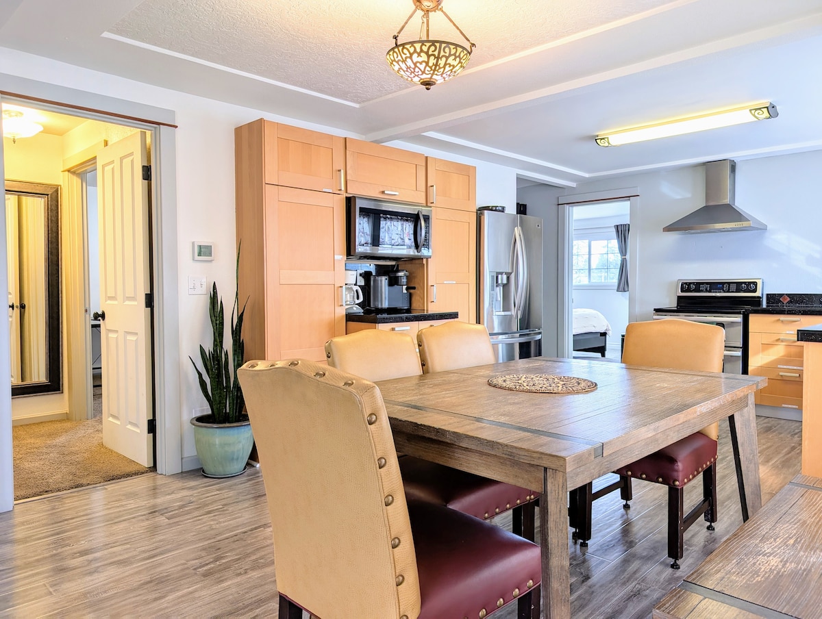 A spacious dining area features a large wooden table surrounded by upholstered chairs. Kitchen appliances, including a microwave and refrigerator, are visible in the background, along with cabinetry and a nearby plant. The open design allows for easy flow between the dining area and kitchen.
