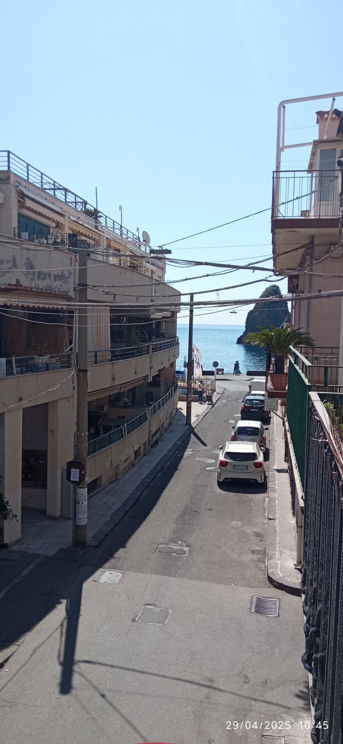 A view of a narrow street lined with buildings on both sides, leading towards the sea. The water is visible at the end of the street, with a small island in the distance. Several parked cars are present along the road.