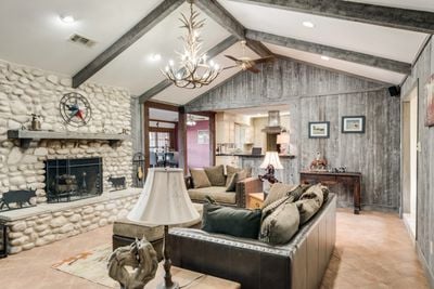 A cozy living area features a stone fireplace with a Texas flag accent, framed by wooden beams overhead. A comfortable leather sofa is adorned with various cushions, accompanied by simple armchairs. Natural light brightens the space, highlighting rustic decor on walls and mantel.