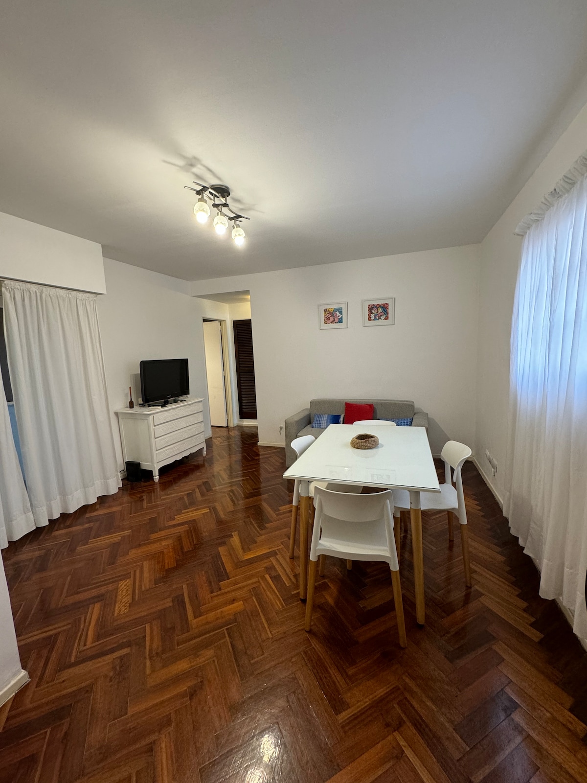 A spacious living area features polished wooden floors and white walls. A simple dining table is surrounded by four chairs, while a sofa sits against one wall. Bright natural light enters through sheer white curtains, and a framed artwork is displayed on the wall.
