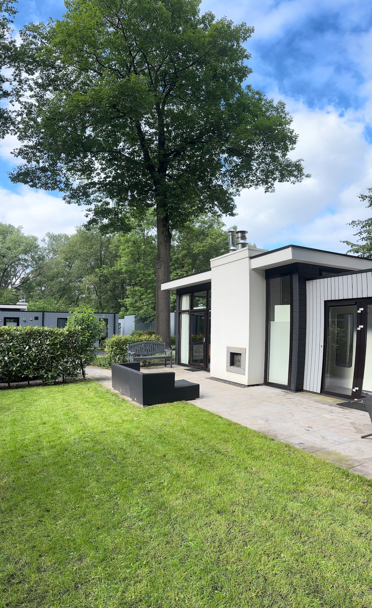 A modern chalet is nestled among lush greenery, with expansive windows allowing natural light to illuminate the space. A comfortable seating arrangement is positioned on a stone patio, enhancing the outdoor living experience. Surrounding grass areas provide ample opportunities for relaxation.