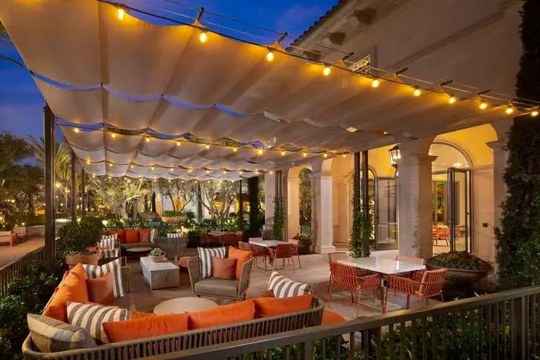 An outdoor lounge area is featured, adorned with comfortable seating that includes sofas and armchairs with orange cushions. Soft lighting hangs overhead, creating a relaxed atmosphere. Lush greenery and garden elements surround the space, offering a serene setting for gatherings.
