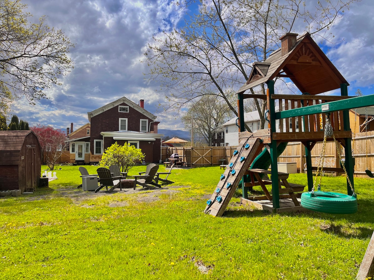 A spacious backyard is showcased with a wooden playset featuring a slide and swings. Adirondack chairs surround a fire pit on the lush green lawn. A shed and a dining area under a canopy are visible in the background, along with the house framed by trees.