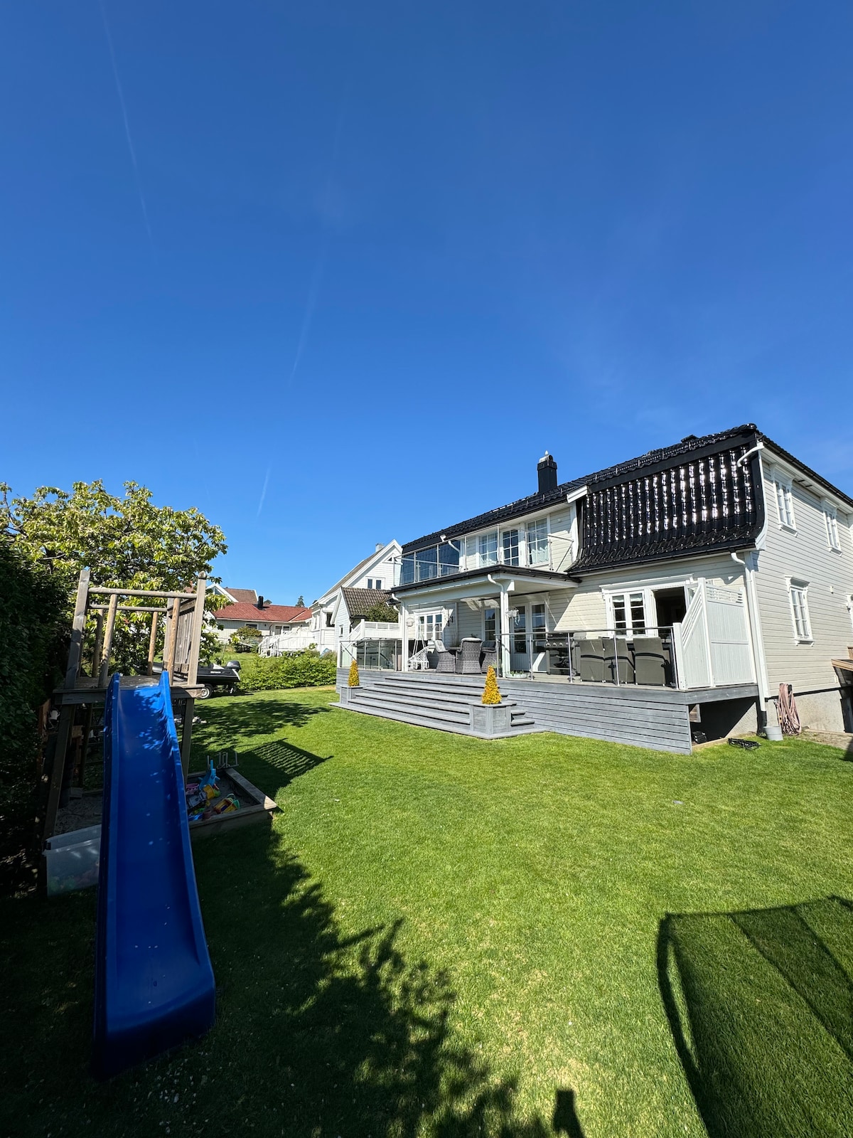 A well-maintained backyard showcases a spacious lawn with a slide and play structure. The two-story house features a terrace with seating, while a balcony offers views of the garden. A clear blue sky enhances the inviting outdoor space.