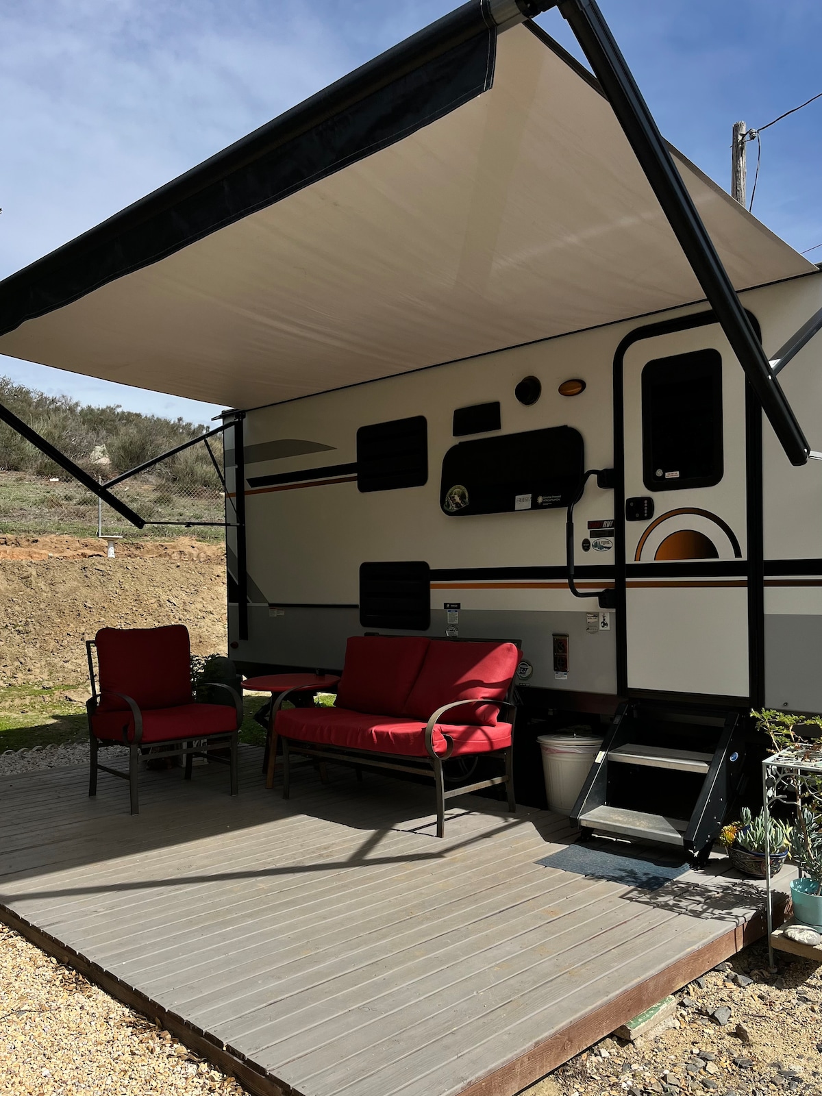 A compact outdoor patio area features a wooden deck shaded by a retractable awning. Two red chairs with metal frames are positioned for seating, accompanied by a small table. Steps lead up to the entrance of the camper, creating a welcoming space for relaxation.