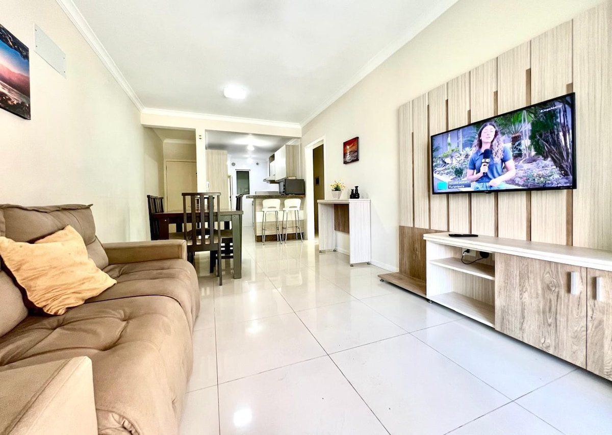 A spacious living area is highlighted by a comfortable sofa with decorative pillows, paired with a dining table that accommodates several guests. A flat-screen television is mounted on a light-colored entertainment unit, reflecting a simple and inviting environment.