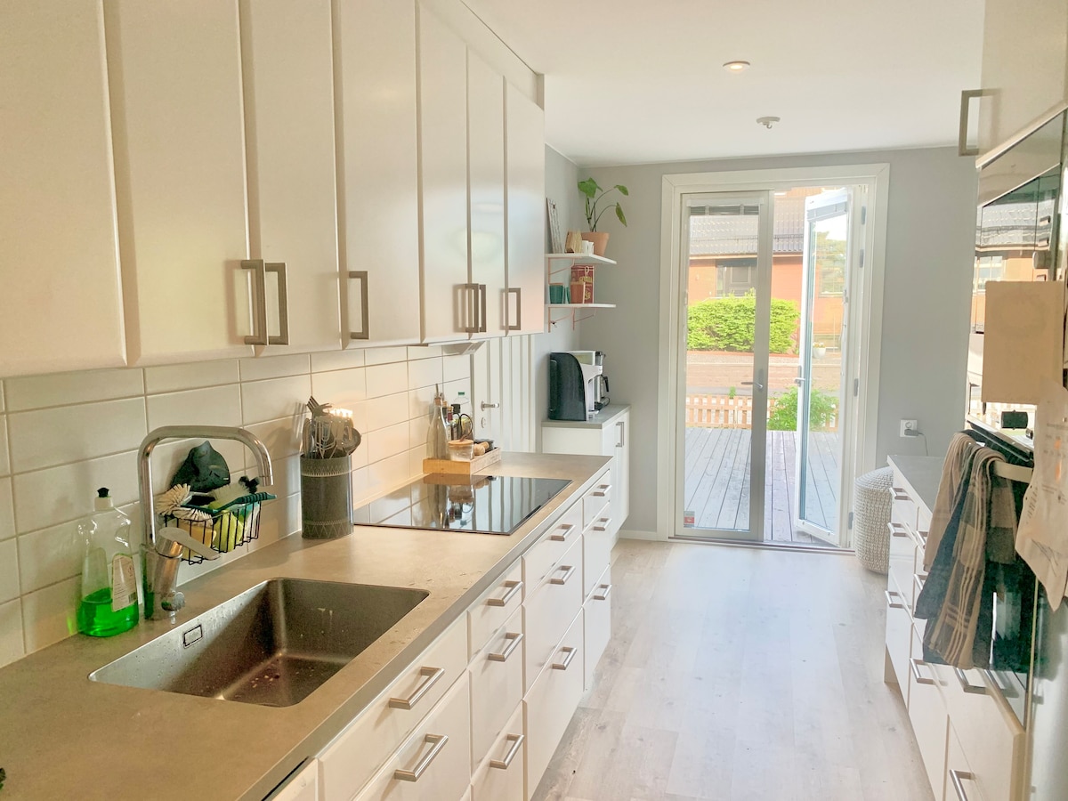 A bright and functional kitchen features white cabinetry with modern hardware. A stainless steel sink is set into a sleek countertop. Natural light fills the space through glass doors that open to the outdoors, showcasing views of the garden.