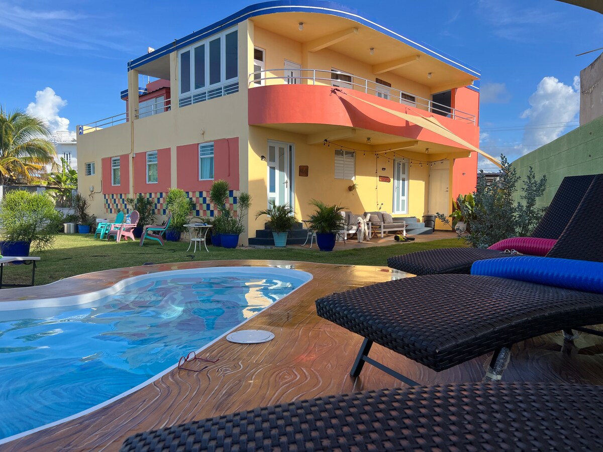 The view showcases a vibrant two-story building with a colorful exterior. A salt-water pool is set in the foreground, surrounded by lounge chairs. Lush greenery and seating areas are present in the spacious yard, inviting relaxation under clear blue skies.