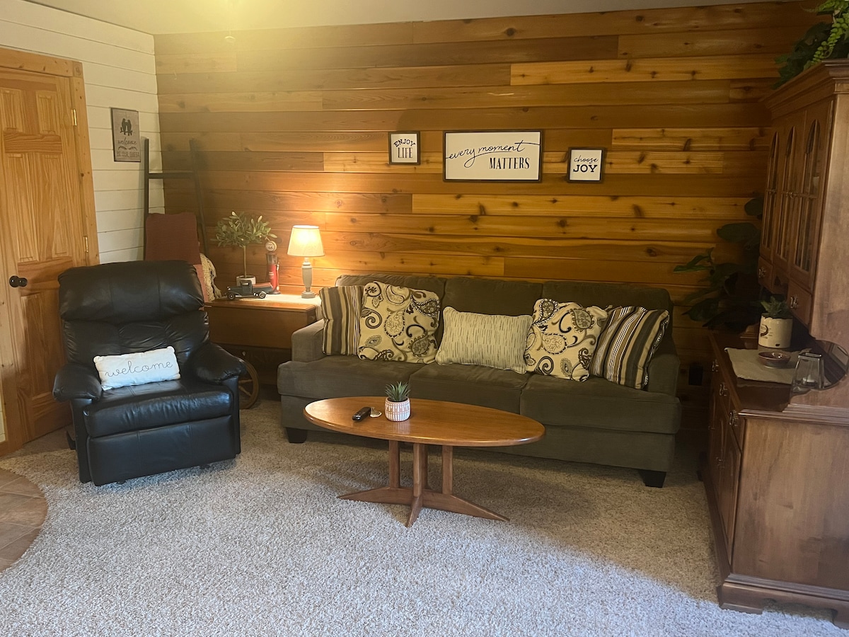 A cozy living room features a plush green sofa adorned with decorative pillows. A wooden coffee table sits in front of the seating area. A recliner is positioned nearby, and a warm light illuminates a rustic wood accent wall, creating a comfortable atmosphere.