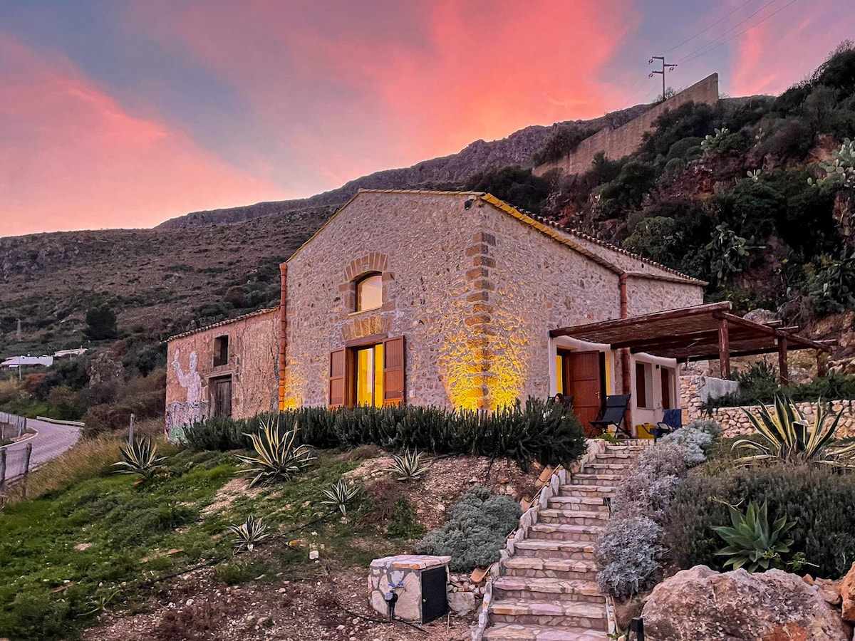 The stone structure features a rustic facade illuminated by evening light. Steps lead to a welcoming entrance framed by greenery and desert plants, with a porch offering shaded seating. A dramatic sunset casts vibrant colors across the sky in the background.