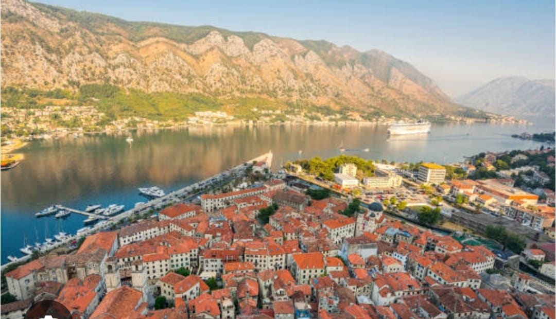 An aerial view captures the terracotta rooftops of Kotor’s Old Town, nestled between the mountains and a calm bay. The waterfront promenade features boats anchored in the marina, while the surrounding landscape displays lush greenery and distant hills under a clear sky.